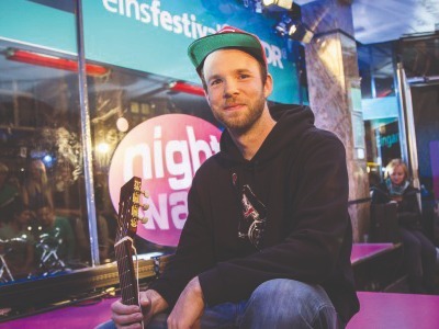Junger Mann mit Bart, Kappe und dunklem Hoodie sitzt mit einer Akustikgitarre vor WDR-Logo und Menschen
