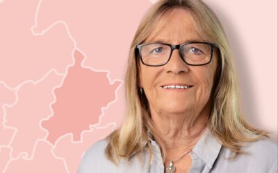 Regina Suett lächelt und trägt Brille vor pinkem Hintergrund mit Landkartenumrissen