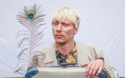 Robert Alan mit blonden Haaren und Bart in Jacke und Hemd, stützt sich auf einen Koffer, daneben eine Pfauenfeder