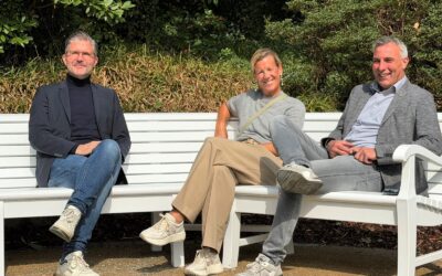 Drei lächelnde Personen sitzen auf einer weißen Parkbank im Freien mit grünen Sträuchern im Hintergrund
