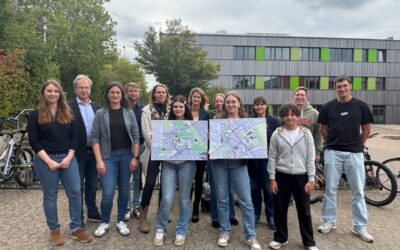Eine Gruppe von Personen steht draußen vor einer Schule, zwei Frauen halten Karten mit Schulwegen in die Kamera