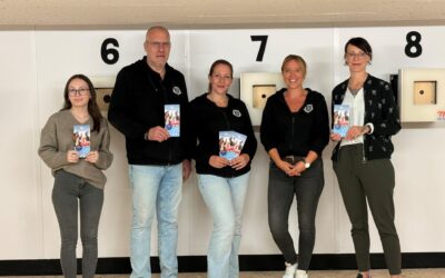 Vier Frauen und ein Mann lächeln und halten Broschüren für „Sporteln & Familienspaß“ in einer Sportanlage