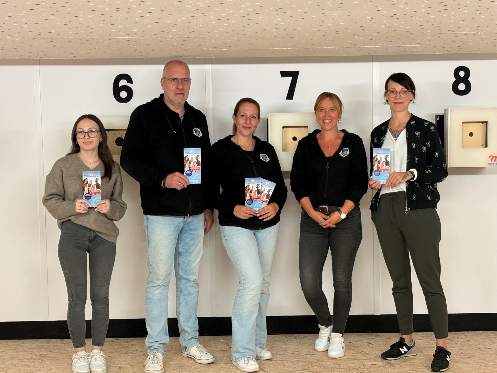 Vier Frauen und ein Mann lächeln und halten Broschüren für „Sporteln & Familienspaß“ in einer Sportanlage