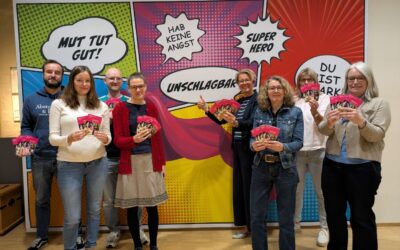 Acht lächelnde Personen präsentieren rosa Programmhefte vor einer Comic-Wand mit Sprechblasen und Motivationssprüchen
