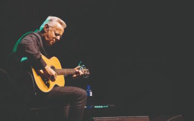 Tommy Emmanuel beugt sich über seine Akustikgitarre und spielt auf einer dunklen Bühne im Scheinwerferlicht