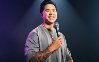 Tutty Tran lächelt, hält ein Mikrofon und zeigt seinen tätowierten Arm unter lila-blauen Scheinwerfern