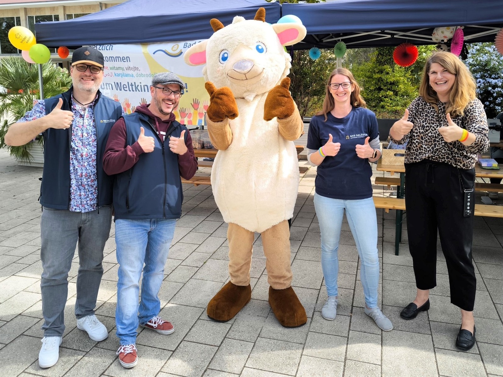 Vier Personen und ein Ziegen-Maskottchen geben Daumen hoch vor einem Weltkindertag-Banner und Zelt
