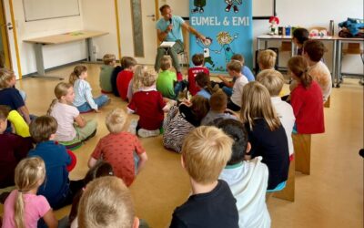 Autor Björn Sassenroth spricht vor sitzenden Grundschülern in einem Raum, hinter ihm ein Banner mit Comicfiguren