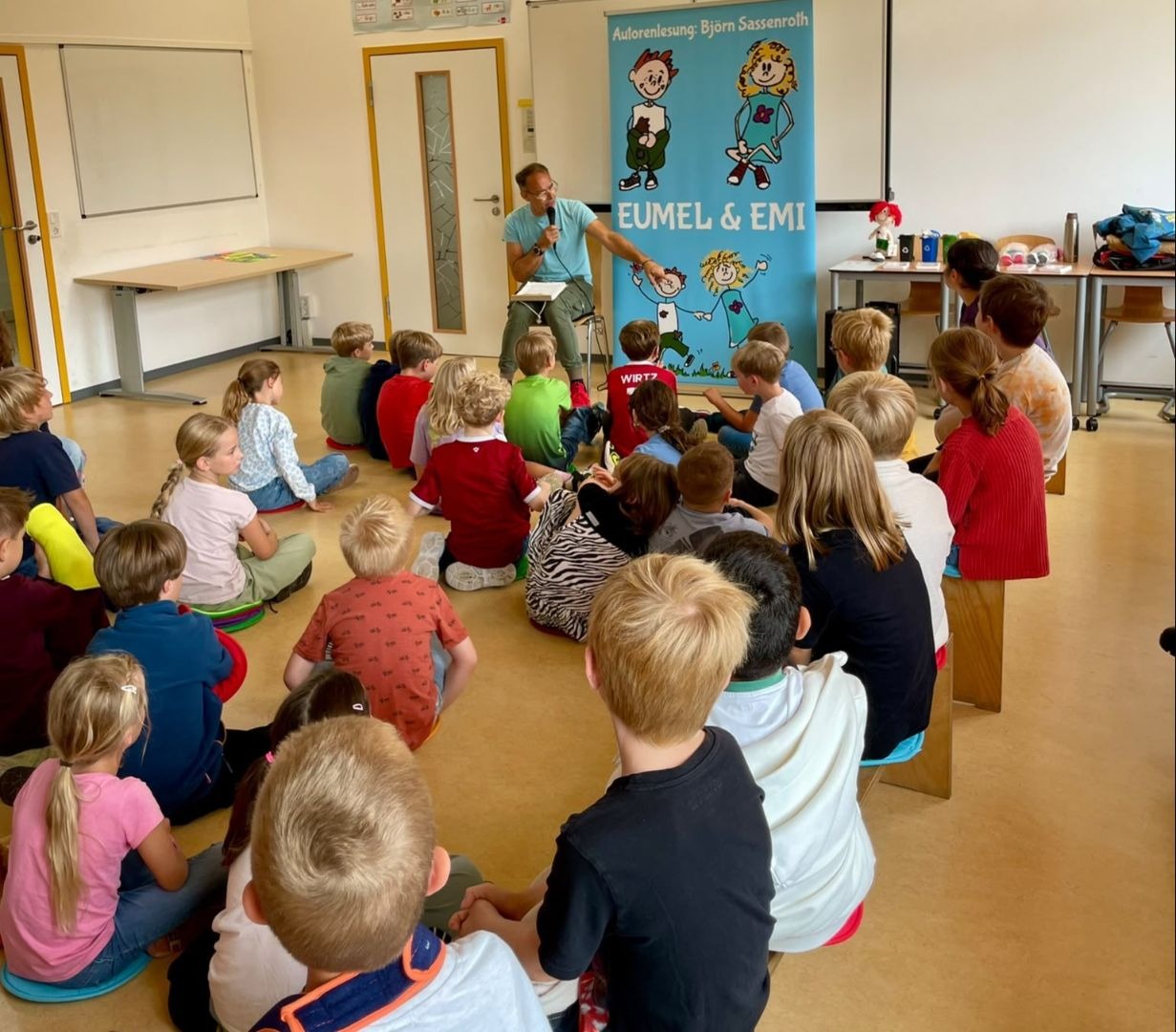 Autor Björn Sassenroth spricht vor sitzenden Grundschülern in einem Raum, hinter ihm ein Banner mit Comicfiguren