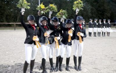 Fünf lächelnde Reiterinnen in Uniform halten Blumensträuße und Siegerschleifen in einer Sandreitbahn hoch