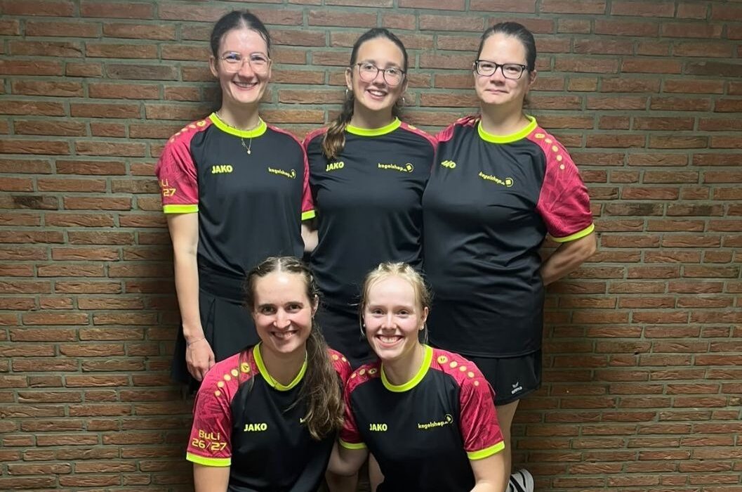 Fünf Frauen in schwarzen Sportshirts und Röcken lächeln, drei stehend und zwei kniend, vor einer Ziegelwand