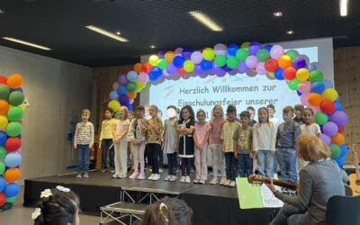 Kinder stehen auf einer Bühne vor einem bunten Ballonbogen und einer Projektion mit "Einschulungsfeier", eine Frau