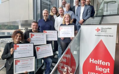 Ein Dutzend Männer und Frauen lächeln auf einer Treppe und halten DRK-Urkunden für Blutspende-Wettbewerbe