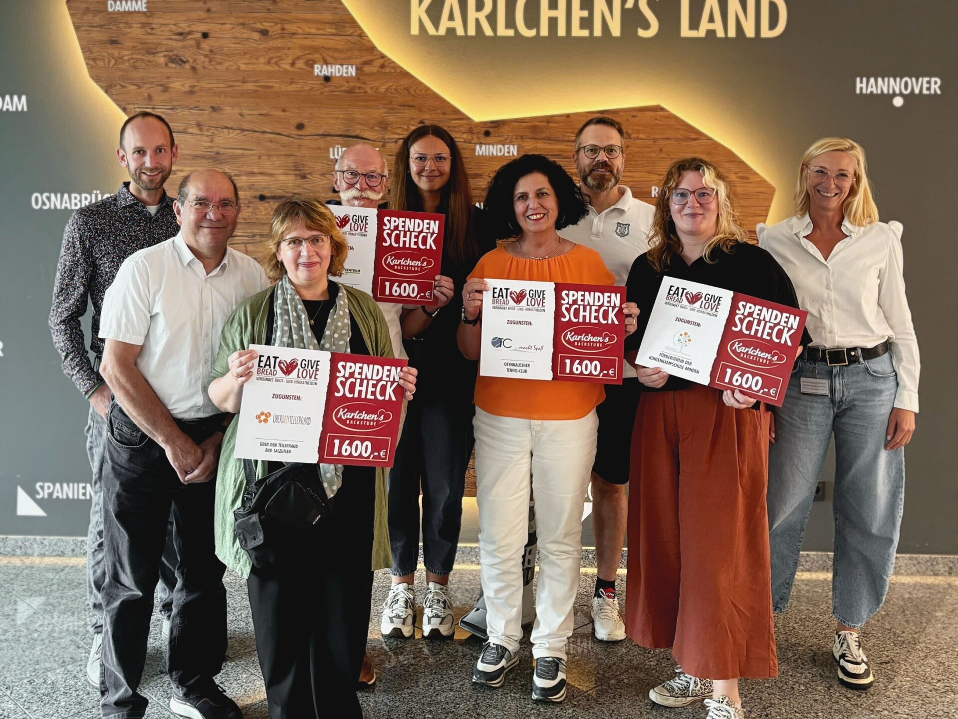 Neun Personen halten rote und weiße Spendenschecks von Karlchen's Backstube über 1600 Euro vor einer Landkarte